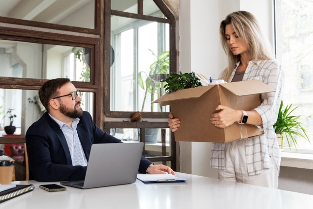 Frau steht mit gepackter Kiste vor einem Mann mit Brille am Laptop